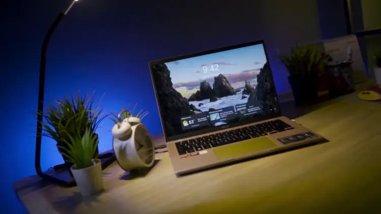 Laptop ASUS Vivobook 14 (M1407) menyala di atas meja kayu dengan latar belakang pencahayaan biru ambient dan tanaman. Gambar unggulan untuk artikel review laptop AI 10 jutaan yang memiliki daya tahan baterai 15 jam.