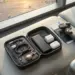 Flat lay koleksi gadget liburan akhir tahun wajib bawa termasuk power bank 20000mAh, charger GaN, gimbal HP, dan AirTag dalam pouch organizer di kursi tunggu bandara saat sunset.
