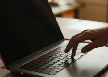 Tangan menekan keyboard laptop dengan layar hitam, memvisualisasikan masalah komputer mati total yang butuh perbaikan.