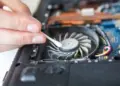Tangan sedang membersihkan debu tebal pada bilah kipas laptop yang berisik, salah satu solusi utama untuk mengatasi masalah Kipas Komputer Berisik dan overheating.