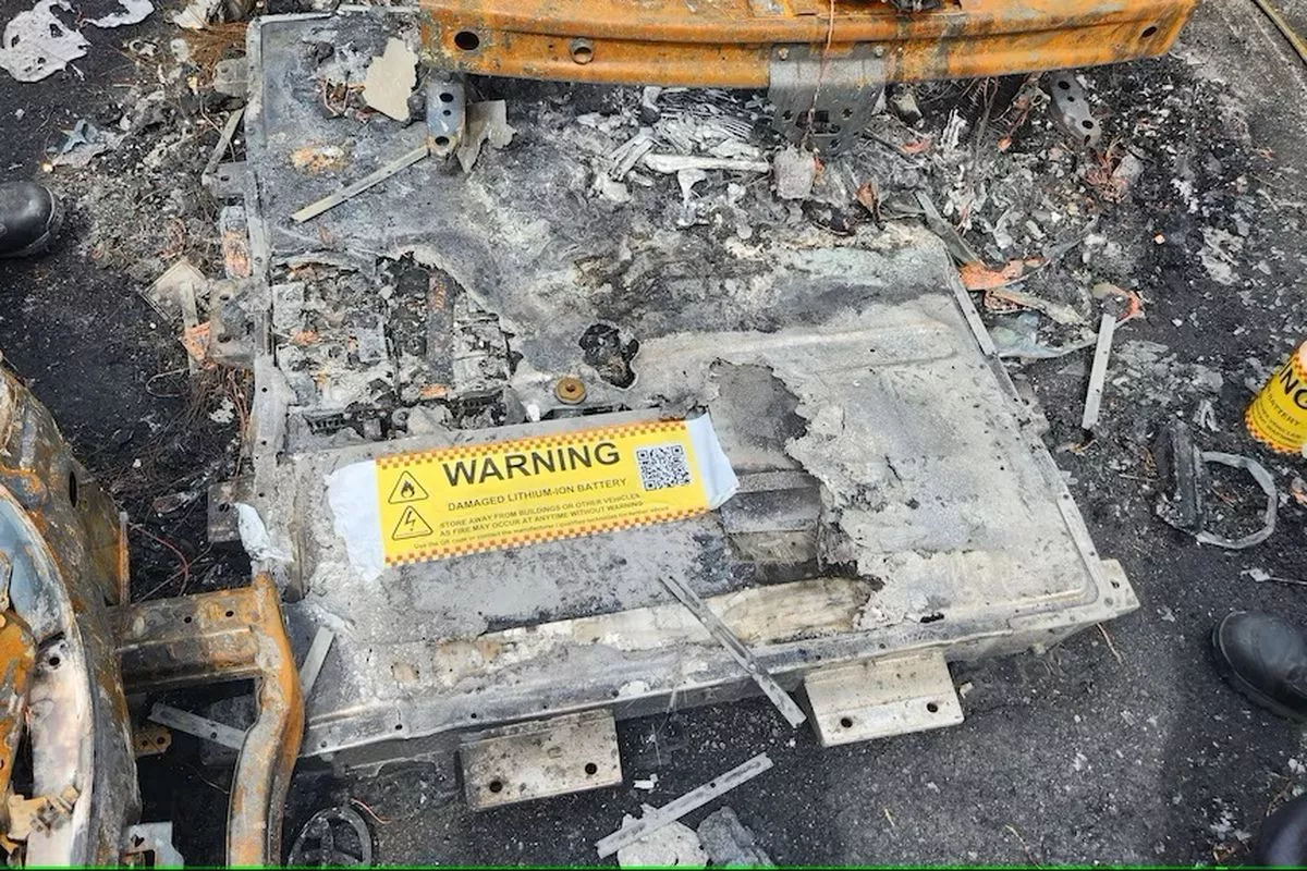 Sisa-sisa baterai lithium-ion yang hancur total akibat fenomena thermal runaway di area terbuka dengan label peringatan kuning.