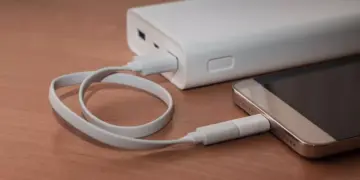 Sebuah smartphone sedang diisi daya menggunakan kabel putih yang terhubung ke Powerbank Terbaik 10.000 mAh di atas meja kayu.