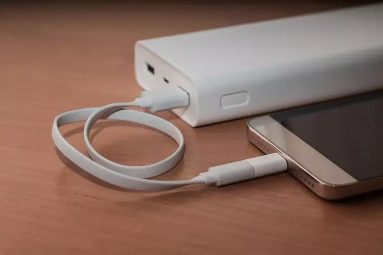 Sebuah smartphone sedang diisi daya menggunakan kabel putih yang terhubung ke Powerbank Terbaik 10.000 mAh di atas meja kayu.