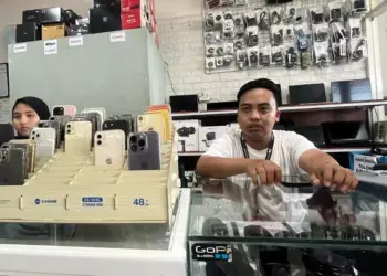 Petugas toko rental iPhone sedang melayani pelanggan dengan jajaran berbagai model iPhone yang siap disewa di etalase.