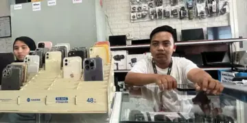 Petugas toko rental iPhone sedang melayani pelanggan dengan jajaran berbagai model iPhone yang siap disewa di etalase.