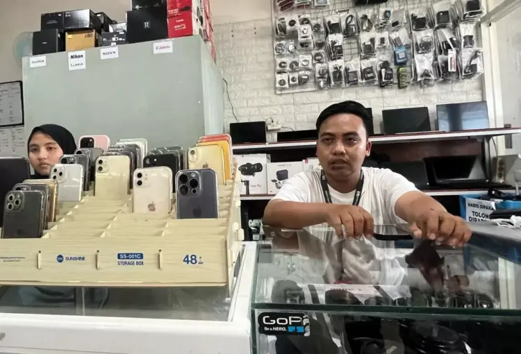 Petugas toko rental iPhone sedang melayani pelanggan dengan jajaran berbagai model iPhone yang siap disewa di etalase.