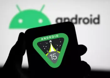 Tangan seseorang memegang smartphone yang menampilkan logo resmi Android 15 berbentuk segitiga dengan ikon roket.