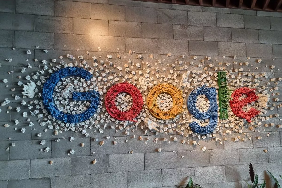 Instalasi seni logo Google di dinding bata yang terbuat dari susunan kerang dan bunga, melambangkan struktur data yang kompleks namun efisien.
