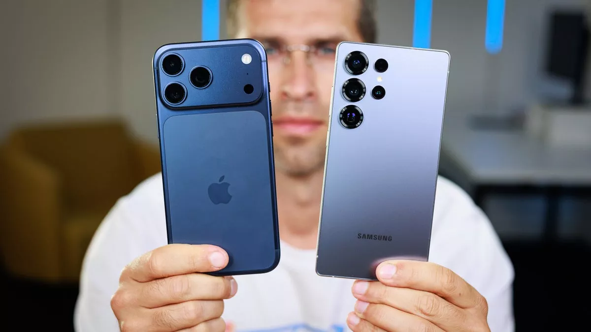 Seorang reviewer memegang Samsung Galaxy S25 Ultra dan iPhone 17 Pro Max warna navy blue untuk perbandingan ukuran.