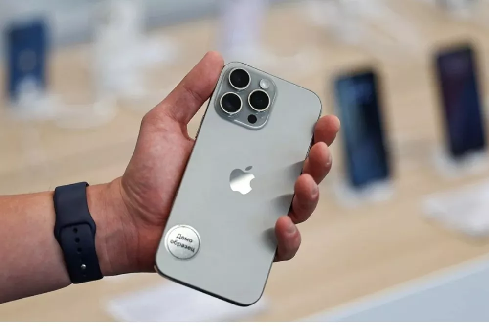 Seseorang mengenakan Apple Watch hitam memegang iPhone 15 Pro berwarna titanium di tangan kanannya, dengan stiker "Demo Unit" di bagian belakang.