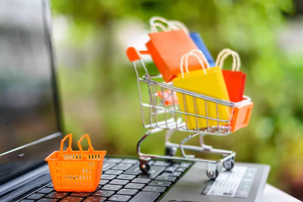 Miniatur keranjang belanja berisi tas belanja warna-warni di atas keyboard laptop, simbol transaksi belanja online.