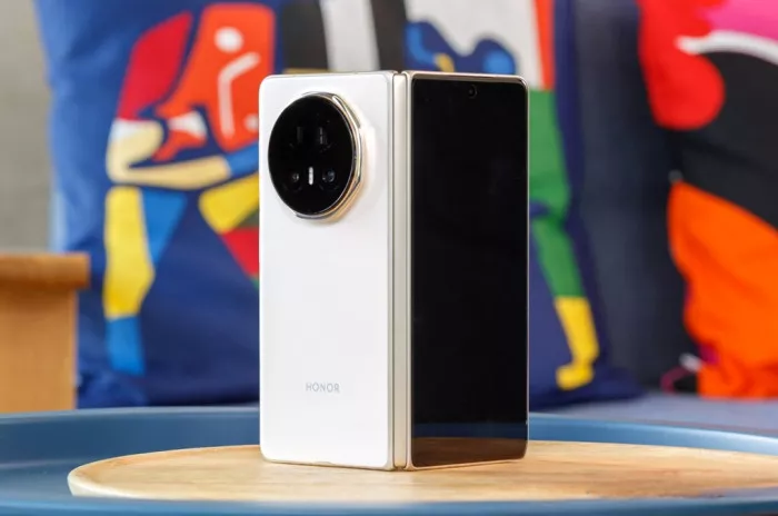 Smartphone lipat Honor Magic V2 warna putih dalam posisi setengah terbuka, diletakkan di atas baki kayu. Kamera belakang berbentuk lingkaran besar.