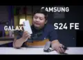 Seorang reviewer pria memegang HP Samsung Galaxy S24 FE putih krem di studio video, menunjuk ponsel tersebut sambil membahas harga Samsung Galaxy S24 FE di tahun 2026 yang turun drastis, menjadikannya pilihan worth it untuk dibeli dengan uang THR Lebaran.