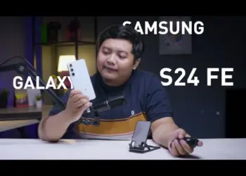 Seorang reviewer pria memegang HP Samsung Galaxy S24 FE putih krem di studio video, menunjuk ponsel tersebut sambil membahas harga Samsung Galaxy S24 FE di tahun 2026 yang turun drastis, menjadikannya pilihan worth it untuk dibeli dengan uang THR Lebaran.