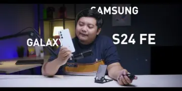 Seorang reviewer pria memegang HP Samsung Galaxy S24 FE putih krem di studio video, menunjuk ponsel tersebut sambil membahas harga Samsung Galaxy S24 FE di tahun 2026 yang turun drastis, menjadikannya pilihan worth it untuk dibeli dengan uang THR Lebaran.