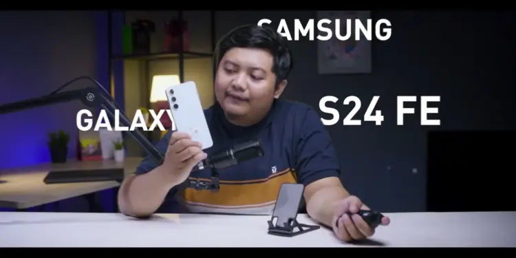 Seorang reviewer pria memegang HP Samsung Galaxy S24 FE putih krem di studio video, menunjuk ponsel tersebut sambil membahas harga Samsung Galaxy S24 FE di tahun 2026 yang turun drastis, menjadikannya pilihan worth it untuk dibeli dengan uang THR Lebaran.
