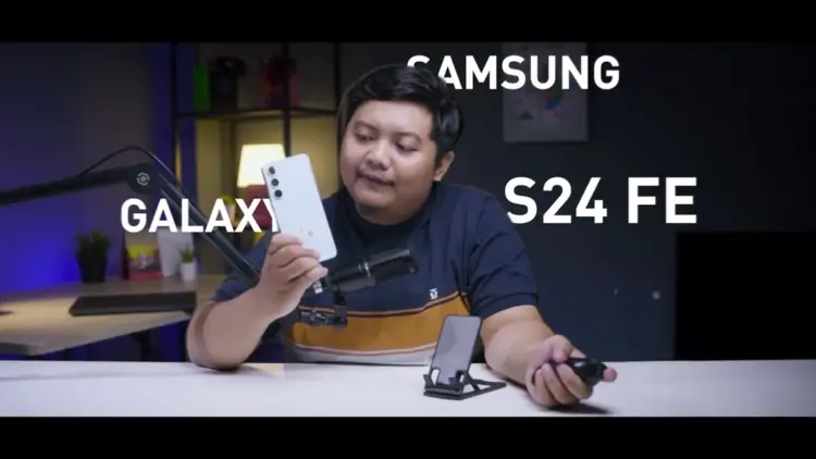 Seorang reviewer pria memegang HP Samsung Galaxy S24 FE putih krem di studio video, menunjuk ponsel tersebut sambil membahas harga Samsung Galaxy S24 FE di tahun 2026 yang turun drastis, menjadikannya pilihan worth it untuk dibeli dengan uang THR Lebaran.