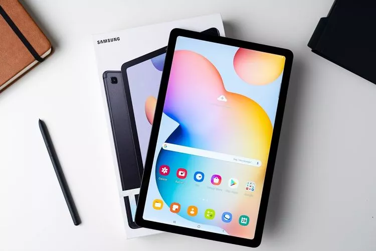 Sebuah tablet Samsung Galaxy Tab S6 Lite berwarna kelabu diletakkan di atas boks kemasannya yang berwarna putih. Di samping kanan tablet, terdapat S Pen berwarna abu-abu gelap, dan di sudut kiri atas ada sebuah buku catatan cokelat. Latar belakangnya adalah permukaan meja putih minimalis. Gambar ini menampilkan salah satu pilihan tablet dengan stylus pen yang siap pakai.