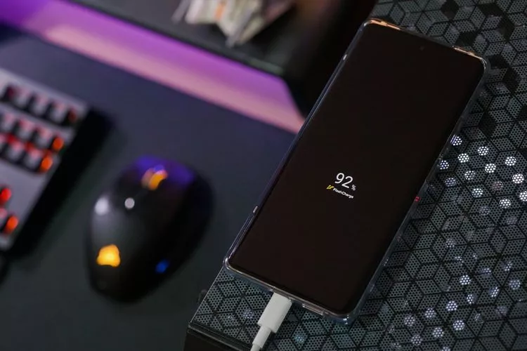Sebuah smartphone sedang diisi daya di atas meja gaming, layar menampilkan persentase baterai 92% dengan ikon Flash Charge.
