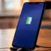 Smartphone berdiri di atas docking charger dengan ikon baterai hijau di tengah layar yang gelap.