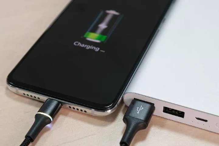 Smartphone terhubung ke power bank menggunakan kabel USB, layar menampilkan animasi pengisian daya bertuliskan "Charging...".
