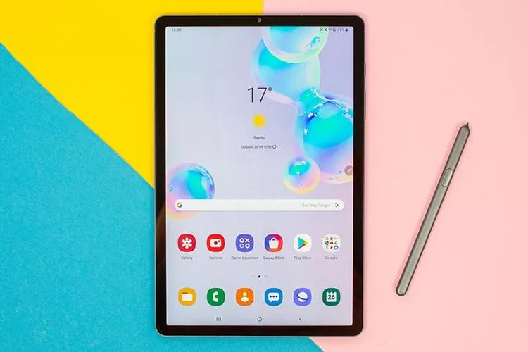 Tampak depan sebuah tablet Samsung Galaxy Tab S6 dengan layar menyala yang menampilkan wallpaper gelembung berwarna-warni dan widget cuaca (17 derajat Celsius). Tablet ini diletakkan di atas permukaan dengan latar belakang color-block setengah kuning cerah di kiri dan setengah merah muda lembut di kanan. S Pen abu-abu diletakkan secara vertikal di sisi kanan tablet, mewakili tablet dengan stylus pen.