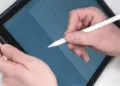 Gambar close-up (jarak dekat) tangan seseorang mengenakan kemeja putih sedang memegang dan menggunakan Apple Pencil generasi pertama berwarna putih pada layar iPad. Di layar iPad yang sedikit dimiringkan, terlihat grid atau grafik berwarna biru. Gambar ini menunjukkan penggunaan langsung salah satu opsi tablet dengan stylus pen.