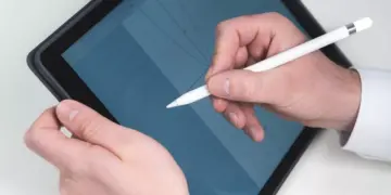 Gambar close-up (jarak dekat) tangan seseorang mengenakan kemeja putih sedang memegang dan menggunakan Apple Pencil generasi pertama berwarna putih pada layar iPad. Di layar iPad yang sedikit dimiringkan, terlihat grid atau grafik berwarna biru. Gambar ini menunjukkan penggunaan langsung salah satu opsi tablet dengan stylus pen.