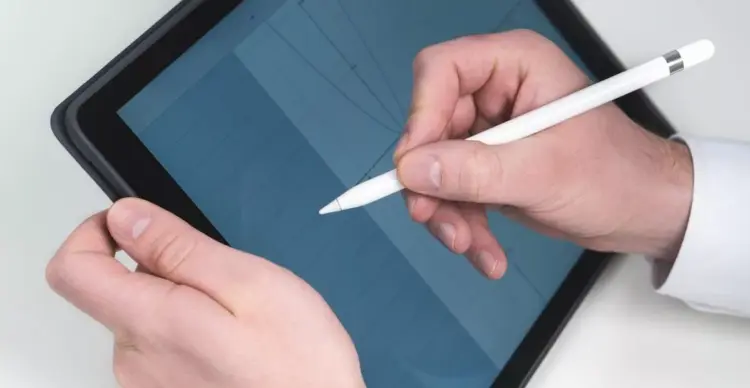 Gambar close-up (jarak dekat) tangan seseorang mengenakan kemeja putih sedang memegang dan menggunakan Apple Pencil generasi pertama berwarna putih pada layar iPad. Di layar iPad yang sedikit dimiringkan, terlihat grid atau grafik berwarna biru. Gambar ini menunjukkan penggunaan langsung salah satu opsi tablet dengan stylus pen.