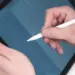 Gambar close-up (jarak dekat) tangan seseorang mengenakan kemeja putih sedang memegang dan menggunakan Apple Pencil generasi pertama berwarna putih pada layar iPad. Di layar iPad yang sedikit dimiringkan, terlihat grid atau grafik berwarna biru. Gambar ini menunjukkan penggunaan langsung salah satu opsi tablet dengan stylus pen.