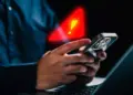 Seseorang memegang smartphone di depan laptop dengan simbol peringatan segitiga merah bercahaya dan tanda seru kuning besar yang melayang di atasnya, melambangkan peringatan infeksi Spyware Morpheus.