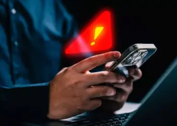 Seseorang memegang smartphone di depan laptop dengan simbol peringatan segitiga merah bercahaya dan tanda seru kuning besar yang melayang di atasnya, melambangkan peringatan infeksi Spyware Morpheus.