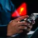 Seseorang memegang smartphone di depan laptop dengan simbol peringatan segitiga merah bercahaya dan tanda seru kuning besar yang melayang di atasnya, melambangkan peringatan infeksi Spyware Morpheus.
