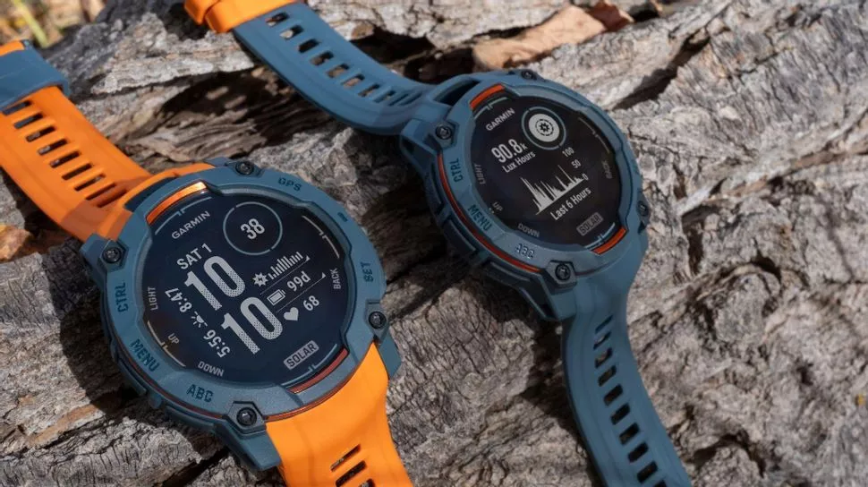 Dua unit Garmin Instinct 3 dengan kombinasi warna strap oranye dan biru diletakkan di atas tekstur batang kayu.