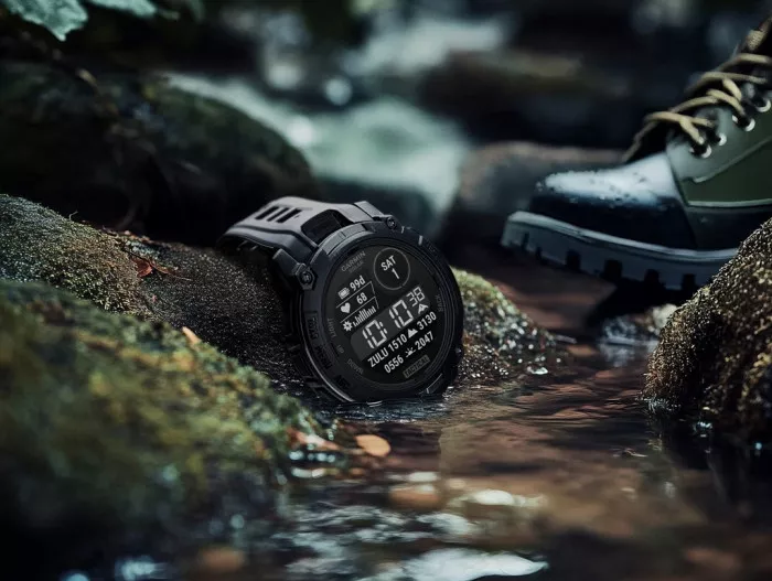 Garmin Instinct 3 edisi Tactical warna hitam diletakkan di atas bebatuan berlumut di aliran sungai.