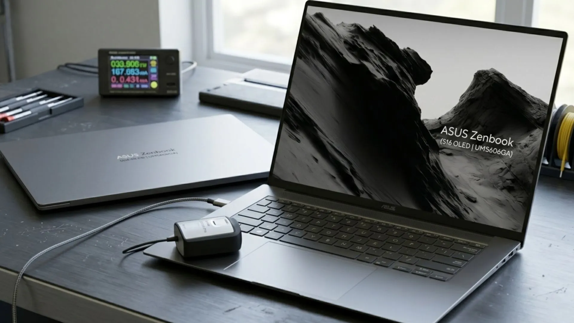 Dua laptop ASUS Zenbook S16 OLED UM5606GA di atas meja kerja, satu terbuka dengan multimeter digital di sampingnya menunjukkan konsumsi daya 33 Watt saat diuji.
