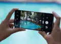 Seseorang memegang smartphone Samsung dengan layar penuh sedang membidik pemandangan kolam renang menggunakan fitur pengunci fokus kamera