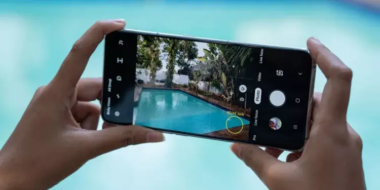 Seseorang memegang smartphone Samsung dengan layar penuh sedang membidik pemandangan kolam renang menggunakan fitur pengunci fokus kamera