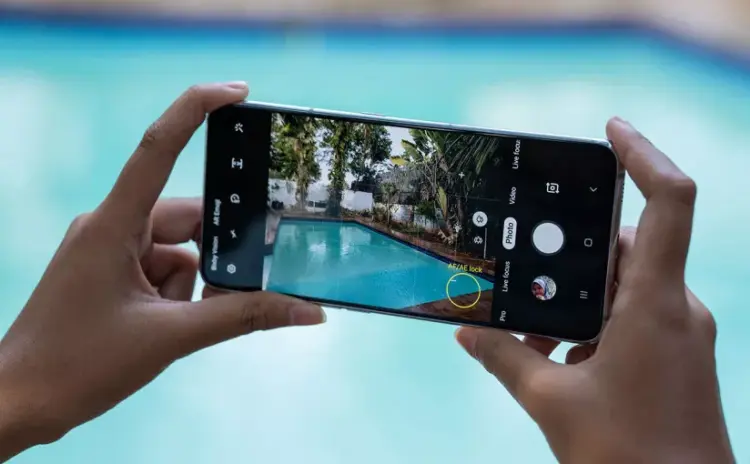 Seseorang memegang smartphone Samsung dengan layar penuh sedang membidik pemandangan kolam renang menggunakan fitur pengunci fokus kamera