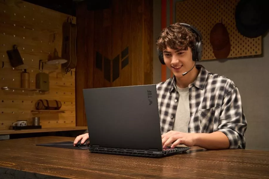 Seorang gamer pria tersenyum menggunakan laptop TUF Gaming F16 hitam dengan headset di ruang bermain.