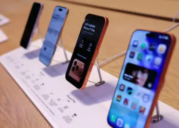 Jajaran unit demo iPhone terbaru yang dipajang di atas meja kayu menggunakan penyangga display khusus toko resmi Apple.