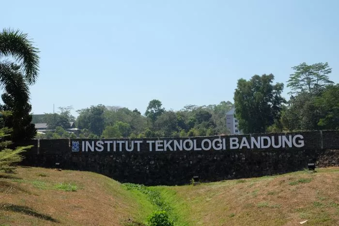 Papan nama ikonik Institut Teknologi Bandung atau ITB dengan latar belakang pepohonan hijau dan langit biru yang cerah.
