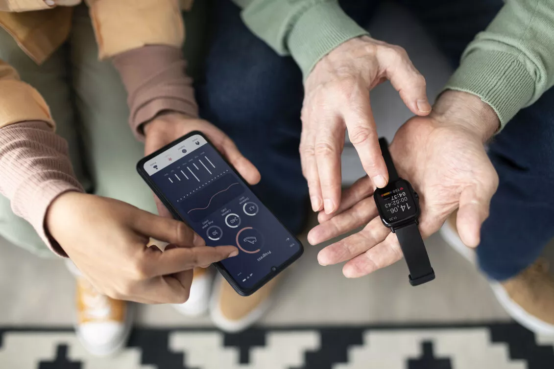 Dua orang sedang mencocokkan data grafik kesehatan antara layar smartphone dan perangkat smartwatch di tangan untuk memantau aktivitas harian.