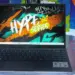Laptop Axioo Hype Series varian Intel Core i3 dengan wallpaper bertuliskan Hype Series diletakkan di atas meja kaca.