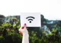 Tangan seseorang memegang kertas putih bergambar logo sinyal internet nirkabel untuk ilustrasi kendala WiFi Limited Access di luar ruangan.