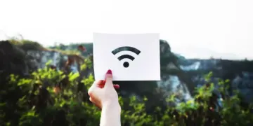 Tangan seseorang memegang kertas putih bergambar logo sinyal internet nirkabel untuk ilustrasi kendala WiFi Limited Access di luar ruangan.