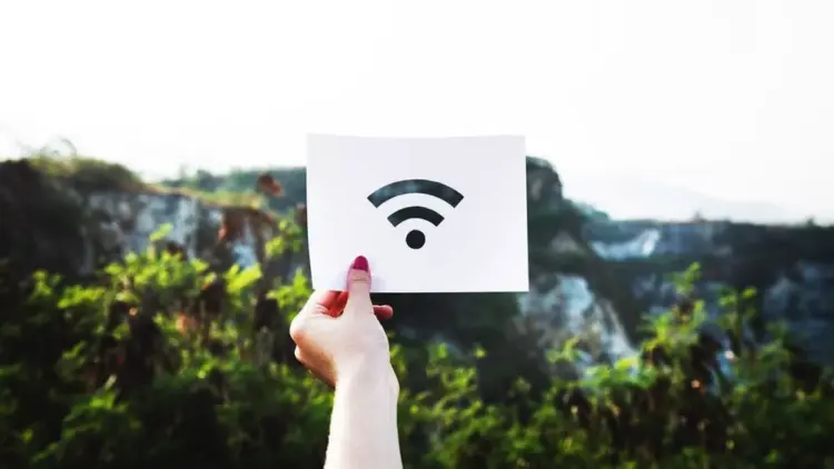 Tangan seseorang memegang kertas putih bergambar logo sinyal internet nirkabel untuk ilustrasi kendala WiFi Limited Access di luar ruangan.