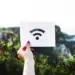 Tangan seseorang memegang kertas putih bergambar logo sinyal internet nirkabel untuk ilustrasi kendala WiFi Limited Access di luar ruangan.