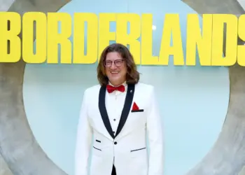 Randy Pitchford mengenakan jas putih tersenyum di depan logo Borderlands, merefleksikan asal usul bocoran aneh Pixel Watch 5.