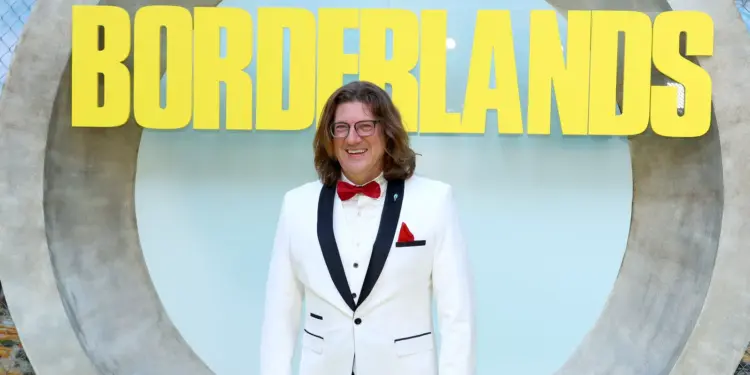Randy Pitchford mengenakan jas putih tersenyum di depan logo Borderlands, merefleksikan asal usul bocoran aneh Pixel Watch 5.
